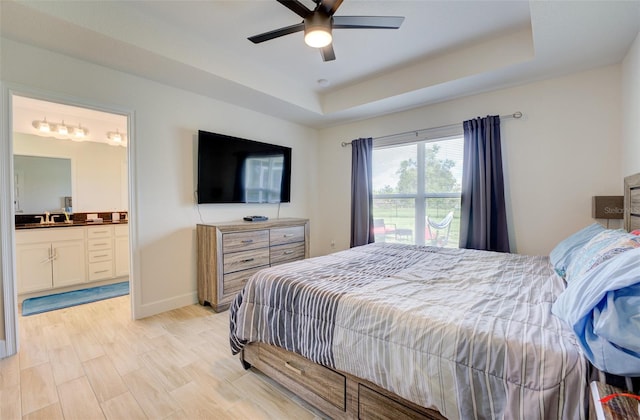 bedroom with light wood-type flooring, ceiling fan, a raised ceiling, ensuite bathroom, and sink