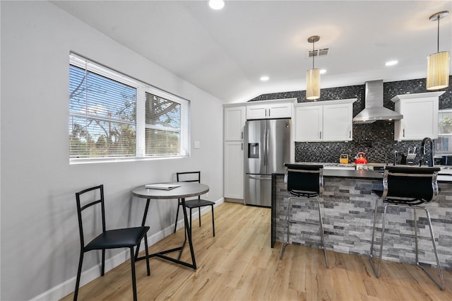 kitchen with decorative light fixtures, white cabinetry, wall chimney range hood, and stainless steel refrigerator with ice dispenser