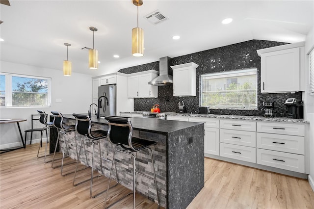 kitchen featuring pendant lighting, wall chimney range hood, white cabinets, a breakfast bar area, and a kitchen island with sink