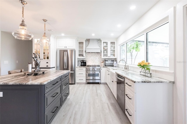 kitchen with white cabinets, hanging light fixtures, appliances with stainless steel finishes, glass insert cabinets, and custom range hood