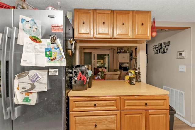 kitchen featuring a textured ceiling, visible vents, light countertops, freestanding refrigerator, and brown cabinets