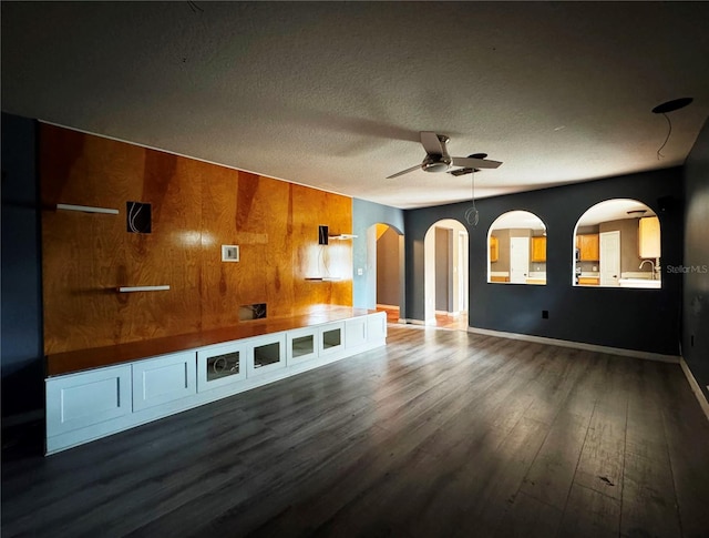 unfurnished living room with arched walkways, dark wood-type flooring, a ceiling fan, a textured ceiling, and wooden walls