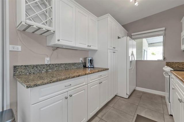 kitchen with light tile patterned floors, white cabinetry, dark stone counters, white appliances, and baseboards