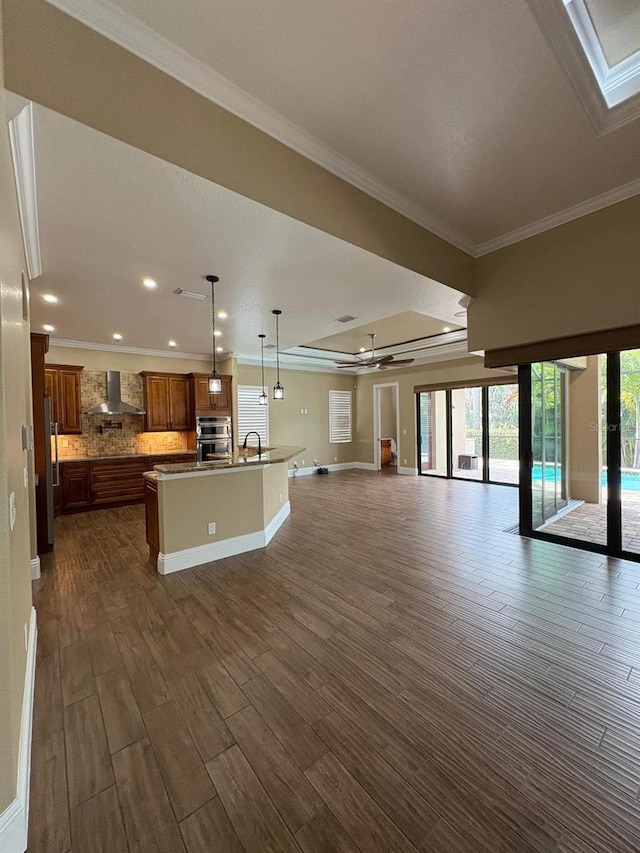 kitchen featuring a center island with sink, brown cabinetry, wall chimney exhaust hood, open floor plan, and pendant lighting