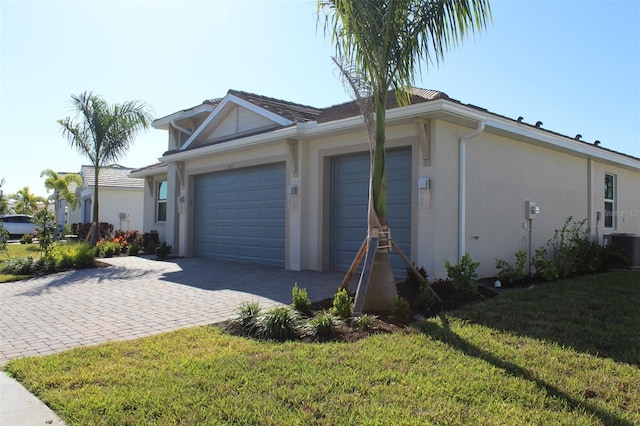 view of side of property featuring decorative driveway, stucco siding, central air condition unit, a lawn, and a garage