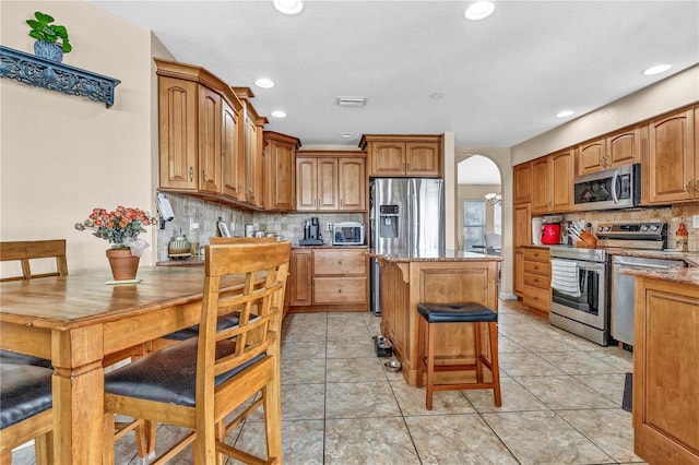 kitchen featuring arched walkways, a breakfast bar area, a kitchen island, appliances with stainless steel finishes, and brown cabinetry