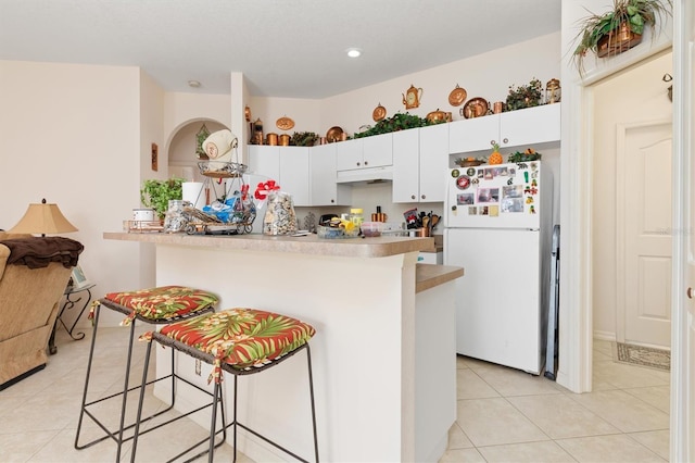 kitchen with light tile patterned flooring, under cabinet range hood, white cabinetry, a kitchen breakfast bar, and freestanding refrigerator