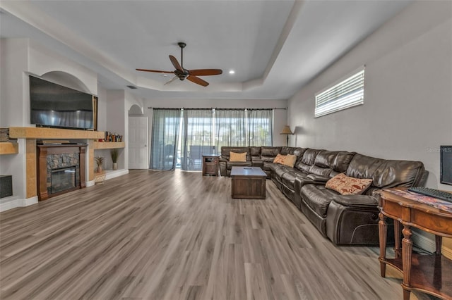 living room featuring a stone fireplace, a tray ceiling, wood finished floors, and a wealth of natural light