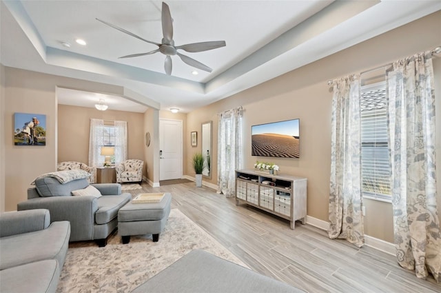 living room with light wood-type flooring, baseboards, a raised ceiling, and a ceiling fan