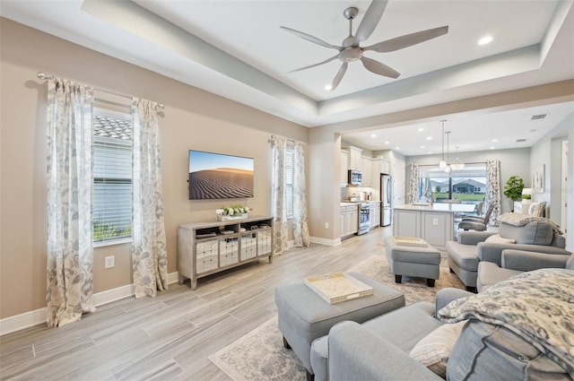 living area with light wood-type flooring, baseboards, a tray ceiling, and a ceiling fan