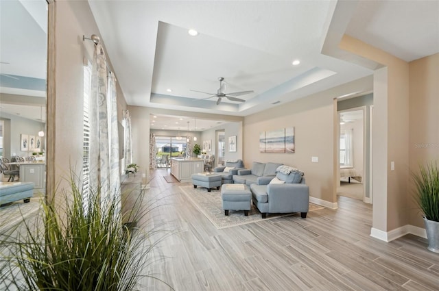living room with ceiling fan, a tray ceiling, baseboards, light wood-style flooring, and recessed lighting