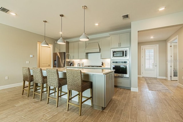 kitchen featuring a center island with sink, gray cabinets, light countertops, appliances with stainless steel finishes, and premium range hood