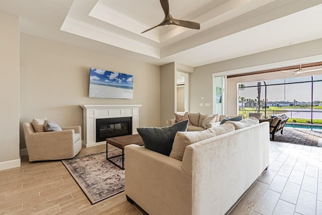 living area featuring light wood finished floors, a raised ceiling, a glass covered fireplace, a sunroom, and ceiling fan