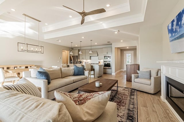 living area with light wood-style flooring, ceiling fan with notable chandelier, ornamental molding, a raised ceiling, and a glass covered fireplace