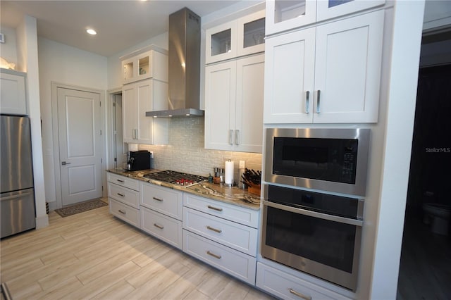 kitchen featuring stainless steel appliances, backsplash, stone countertops, light wood-type flooring, and wall chimney exhaust hood