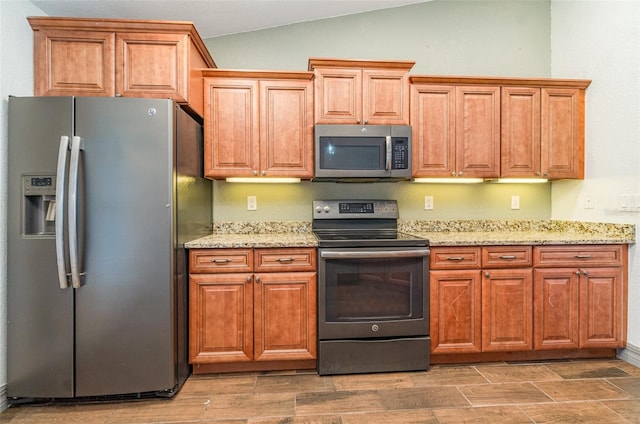 kitchen featuring lofted ceiling, appliances with stainless steel finishes, brown cabinetry, wood tiled floor, and light stone countertops