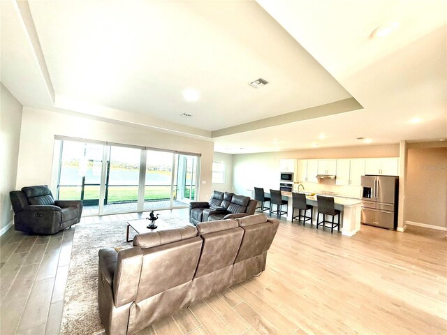 living room with light wood-type flooring, visible vents, a tray ceiling, and baseboards