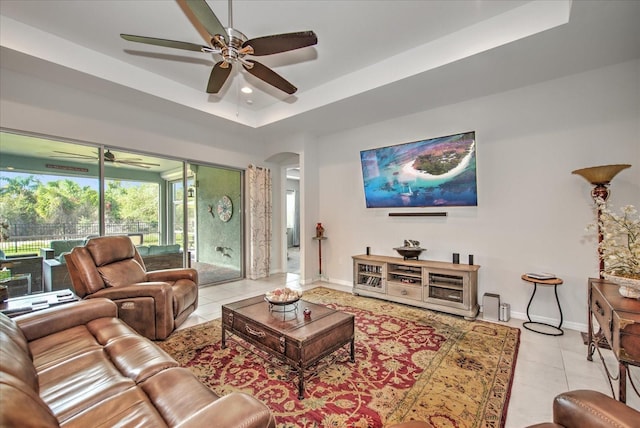 living room featuring arched walkways, light tile patterned floors, a ceiling fan, baseboards, and a raised ceiling