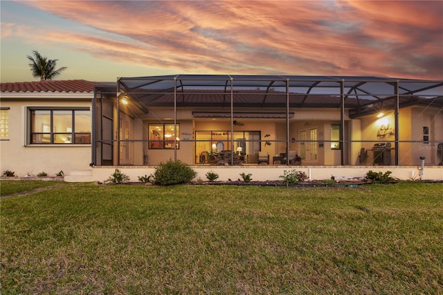 back of house at dusk with stucco siding, a tiled roof, glass enclosure, and a yard