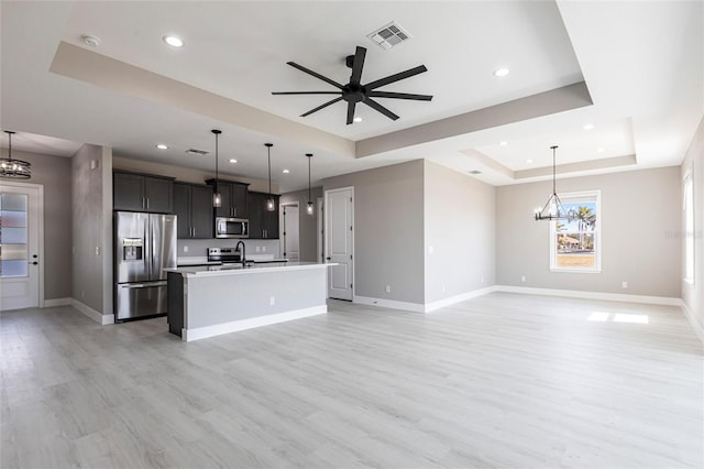 kitchen with a tray ceiling, stainless steel appliances, open floor plan, an island with sink, and ceiling fan with notable chandelier