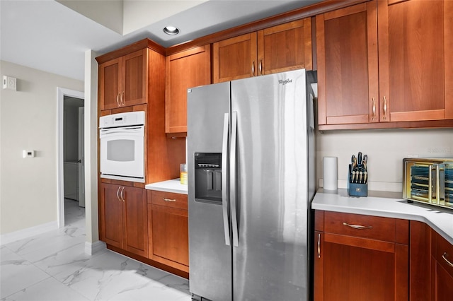 kitchen featuring brown cabinetry, light countertops, stainless steel refrigerator with ice dispenser, white oven, and marble finish floor