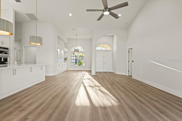 unfurnished living room with a ceiling fan, visible vents, baseboards, light wood-style flooring, and a towering ceiling