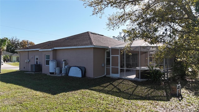 rear view of house with glass enclosure, central AC unit, roof with shingles, a yard, and stucco siding