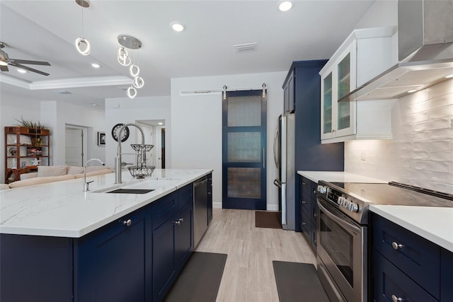 kitchen featuring blue cabinetry, wall chimney range hood, a barn door, decorative backsplash, and stainless steel appliances