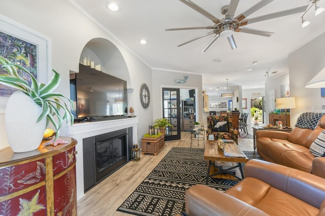 living area featuring light wood-type flooring, a ceiling fan, a glass covered fireplace, recessed lighting, and crown molding