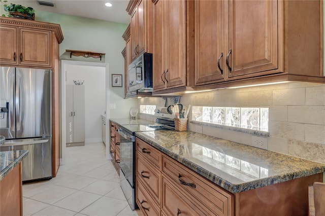 kitchen with light tile patterned floors, decorative backsplash, dark stone countertops, appliances with stainless steel finishes, and brown cabinetry