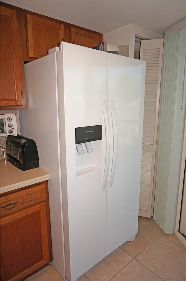 kitchen featuring white fridge with ice dispenser, light tile patterned flooring, brown cabinetry, and light countertops