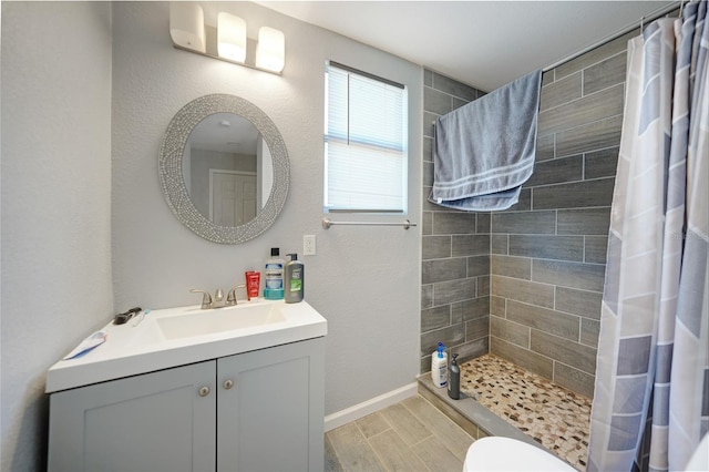 bathroom featuring baseboards, wood tiled floor, toilet, tiled shower, and vanity