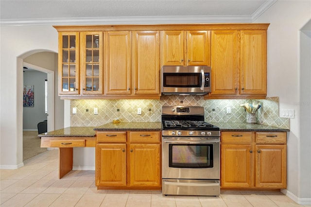 kitchen featuring glass insert cabinets, arched walkways, ornamental molding, dark stone counters, and stainless steel appliances