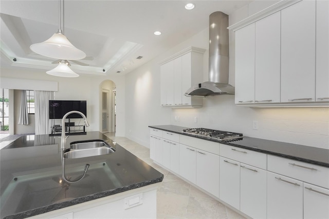 kitchen featuring arched walkways, a sink, wall chimney range hood, a tray ceiling, and stainless steel gas cooktop