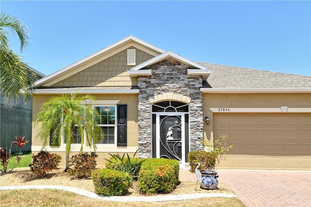 view of front of property featuring stone siding, an attached garage, a shingled roof, stucco siding, and decorative driveway