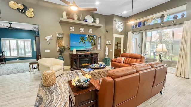 living room featuring a high ceiling, light wood finished floors, a ceiling fan, baseboards, and a glass covered fireplace