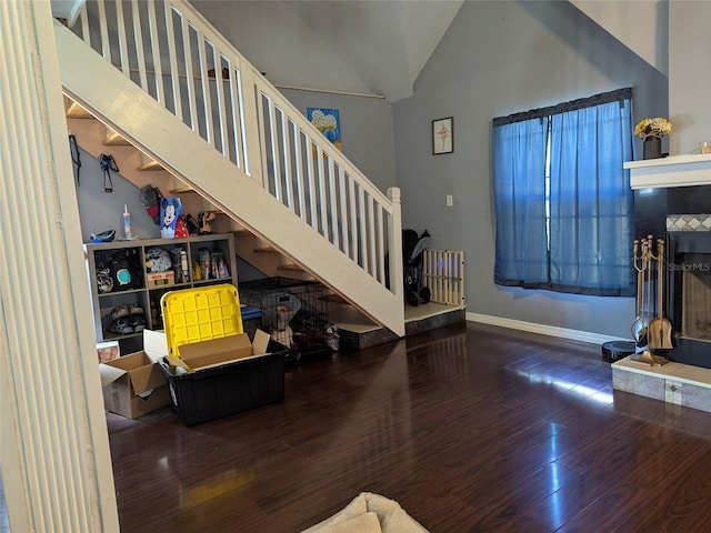 living area featuring high vaulted ceiling, a tiled fireplace, baseboards, wood finished floors, and stairway