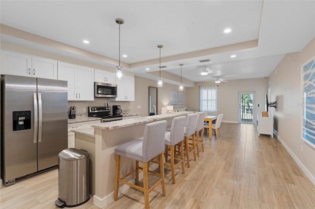 kitchen featuring appliances with stainless steel finishes, a tray ceiling, an island with sink, light wood-type flooring, and recessed lighting