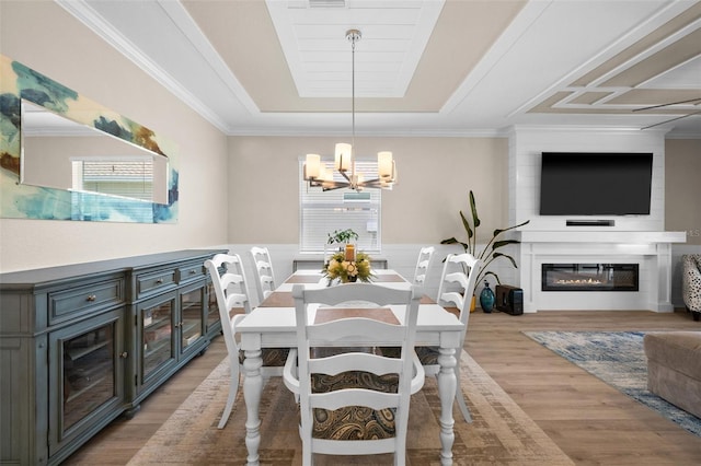 dining room featuring a raised ceiling, a chandelier, a glass covered fireplace, light wood-style flooring, and ornamental molding