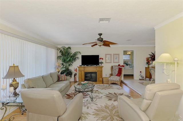 living room featuring visible vents, crown molding, a glass covered fireplace, ceiling fan, and light wood-type flooring