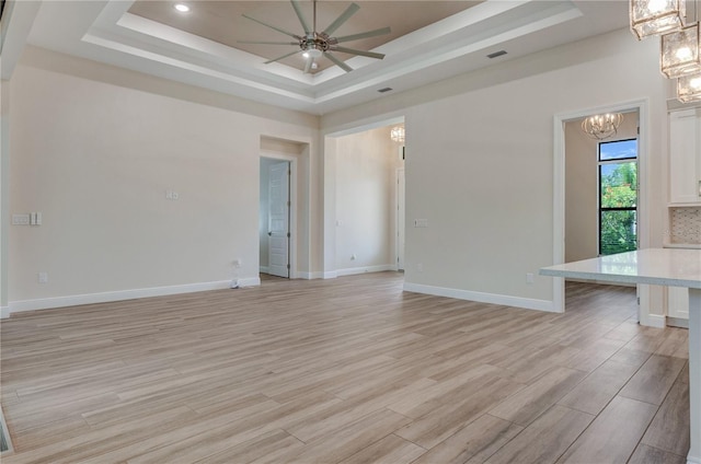 unfurnished living room with a raised ceiling, a chandelier, ceiling fan, light wood-style flooring, and baseboards