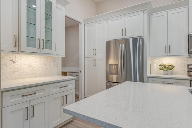 kitchen with stainless steel refrigerator with ice dispenser, glass insert cabinets, light stone counters, and backsplash