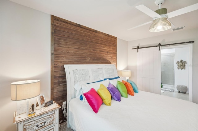 bedroom with a barn door, wooden walls, ceiling fan, ensuite bath, and tile patterned floors