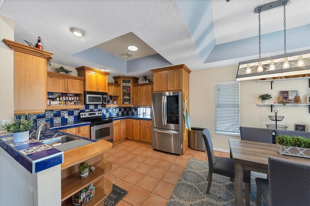 kitchen featuring dark countertops, glass insert cabinets, appliances with stainless steel finishes, a tray ceiling, and a sink