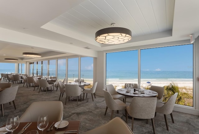 dining space with a water view, a wall of windows, a tray ceiling, and a view of the beach