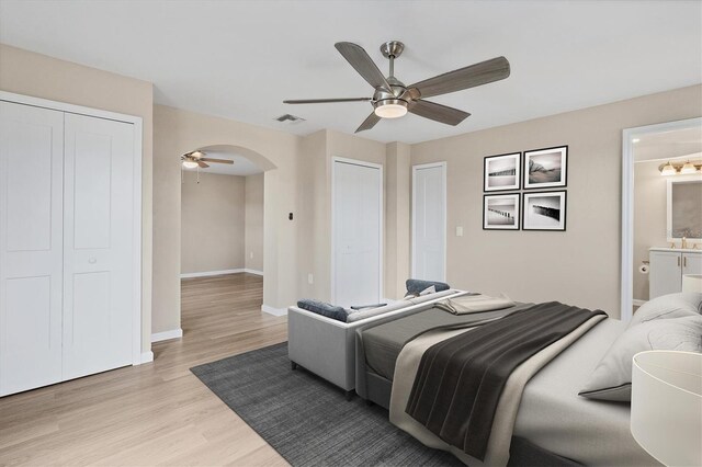 bedroom featuring two closets, ensuite bathroom, light wood-type flooring, and ceiling fan