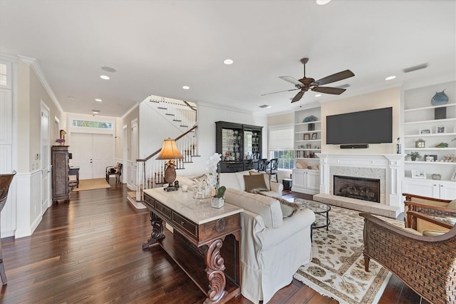 living room featuring dark wood-type flooring, ceiling fan, a premium fireplace, built in shelves, and ornamental molding