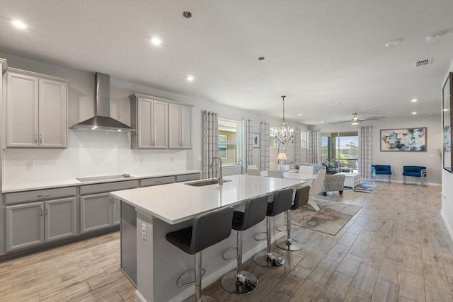 kitchen with ceiling fan with notable chandelier, sink, wall chimney range hood, a center island with sink, and gray cabinets