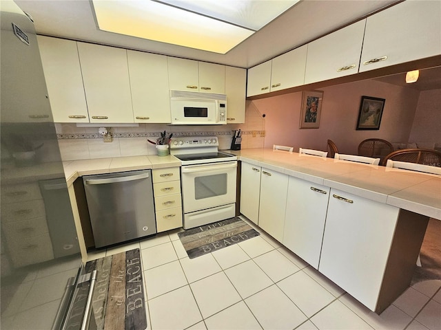 kitchen with backsplash, kitchen peninsula, light tile patterned flooring, white appliances, and white cabinets