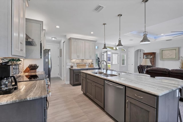 kitchen with sink, dishwasher, wall chimney range hood, light wood-type flooring, and an island with sink